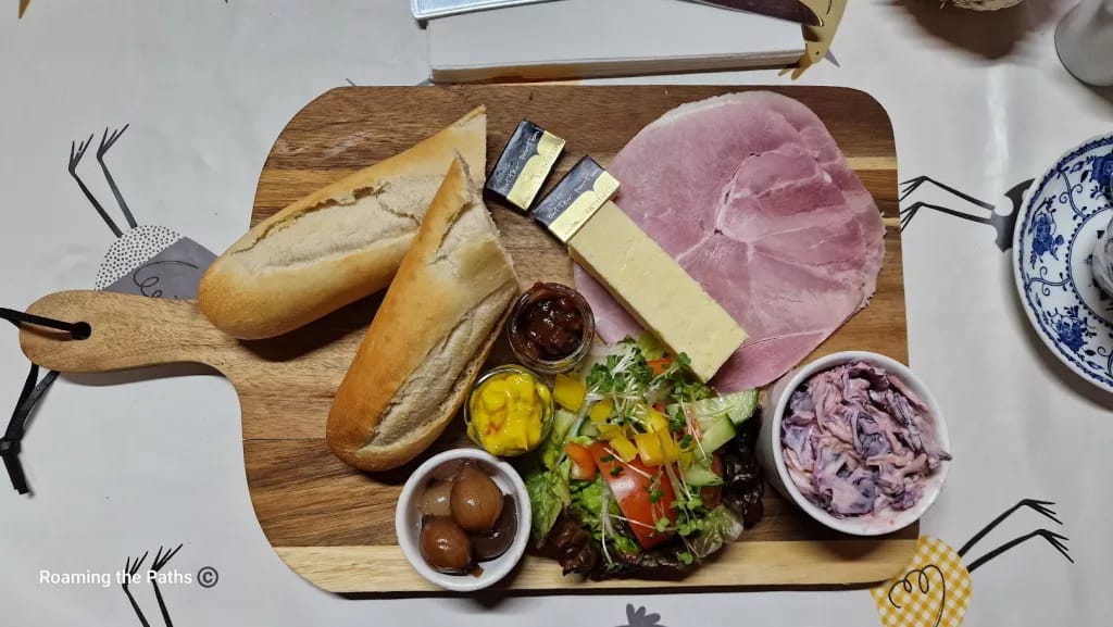 
This image shows a beautifully arranged Ploughman’s Lunch at The Old Forge, displayed on a wooden serving board. The classic British meal includes:

Two baguette halves – Fresh and crusty, perfect for pairing with the cheese and ham.
Thick slice of cheddar cheese – A creamy, yellow wedge of cheddar, a traditional element of the Ploughman’s.
Sliced ham – Generous, thin slices of pink ham, offering a mild, savory flavor.
Condiments and accompaniments:
A small jar of chutney, likely caramelized onion or fruit-based, for a sweet and tangy kick.
A small jar of piccalilli – a bright yellow relish made from pickled vegetables in a mustard sauce, adding a sharp, tangy note.
A small bowl of pickled onions, adding a sour punch.
Mixed side salad – Fresh lettuce, tomatoes, cucumber, and yellow bell pepper with microgreens, bringing vibrant color and crunch.
Creamy coleslaw – A bowl of pinkish coleslaw with red cabbage, adding a creamy and crunchy texture.
The board rests on a farm-themed tablecloth featuring playful chicken illustrations, enhancing the rustic and homely presentation of this classic Ploughman’s Lunch.