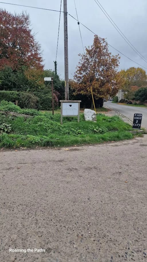 This image shows a subtle roadside entrance to a farm shop and café. A small, understated sign on a wooden frame reads "The Old Forge" with the shop’s logo, blending into the rustic and natural surroundings.

Nearby, a black sandwich board on the ground boldly states “Farm Shop & Café,” making it more noticeable to passing vehicles. The area is surrounded by lush greenery, with mature trees displaying autumnal colors of orange and red. An overhead power pole and country road emphasize the rural location, while the grassy verge and unpaved roadside give it a quiet, countryside feel. This signage invites visitors to explore the hidden gem of the farm shop and café nearby.