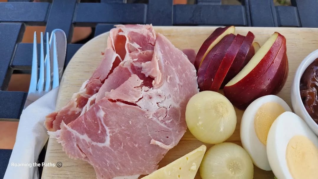 Close-up of a wooden platter featuring thick slices of rustic ham, sliced red apple, pickled onions, and halved boiled eggs. A wedge of cheese and a glimpse of chutney in a ramekin are visible on the side. A napkin-wrapped knife and fork rest on the edge of the board, all set atop a black metal outdoor table.