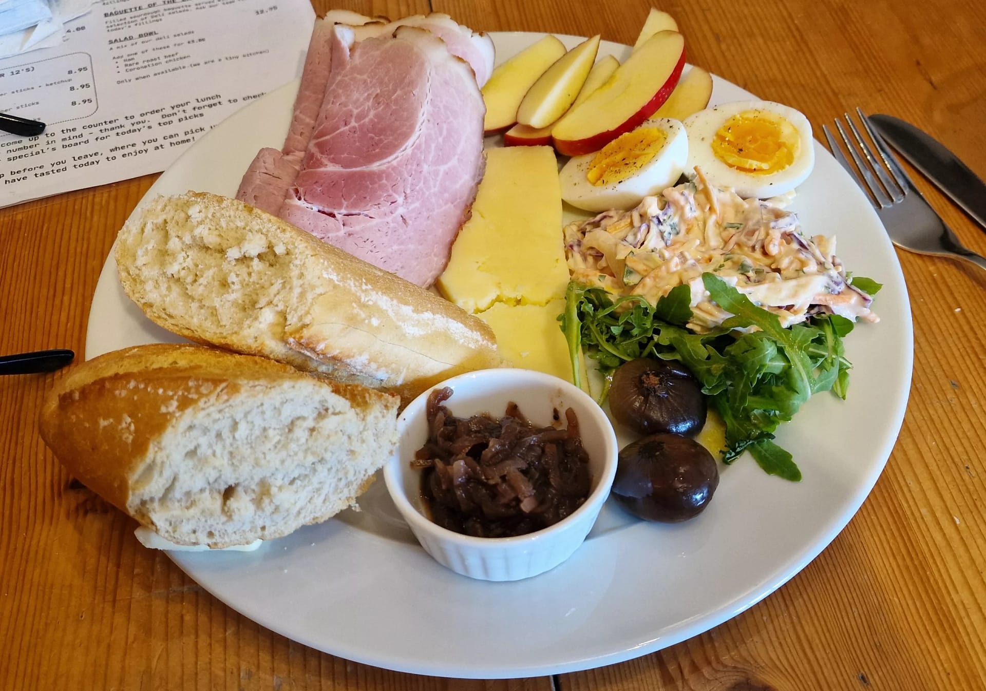 A generously filled Ploughman’s lunch served on a white plate, featuring thick slices of pink ham, a wedge of golden cheddar cheese, and crisp apple slices. Beside them are two halves of a hard-boiled egg, a creamy mound of coleslaw, fresh green salad leaves, and dark pickled onions. At the front sits a crusty baguette, torn open to show its soft inside, alongside a small white ramekin of tangy chutney. The meal is laid out on a wooden table, evoking the hearty, rustic style of a traditional English pub platter.