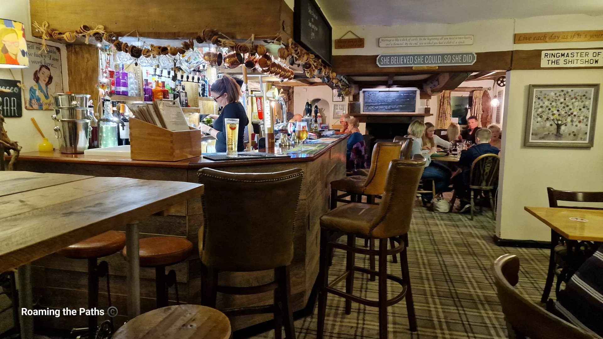 Interior of a cosy country pub showing the bar area with hanging copper mugs, bottles of spirits, and a bartender serving drinks, while diners enjoy meals in a warmly lit seating area beyond.