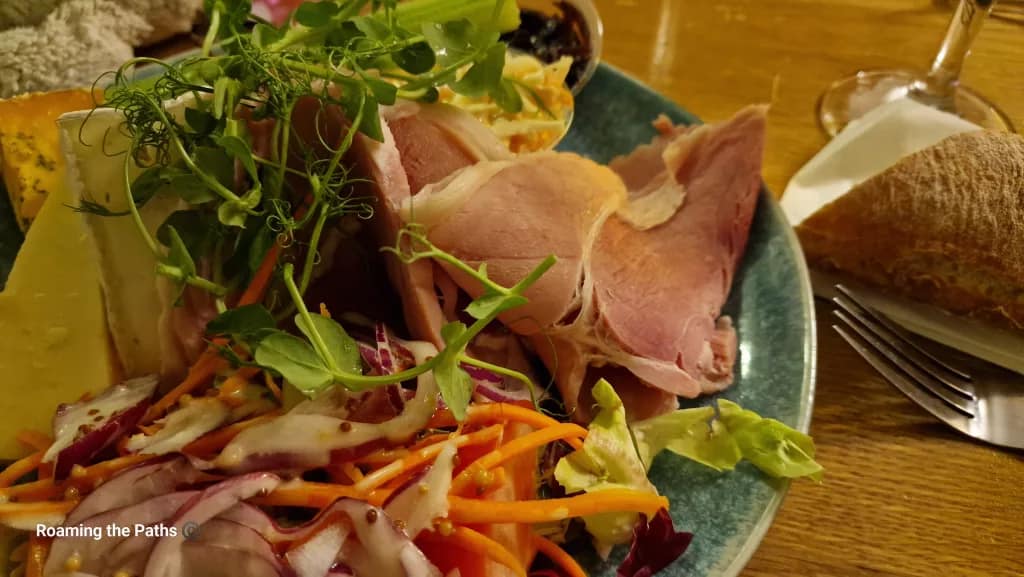 Close-up of a Ploughman’s lunch showing thick slices of ham, Brie and Cheddar cheese, colourful salad with mustard dressing, and pea shoots on a rustic plate.