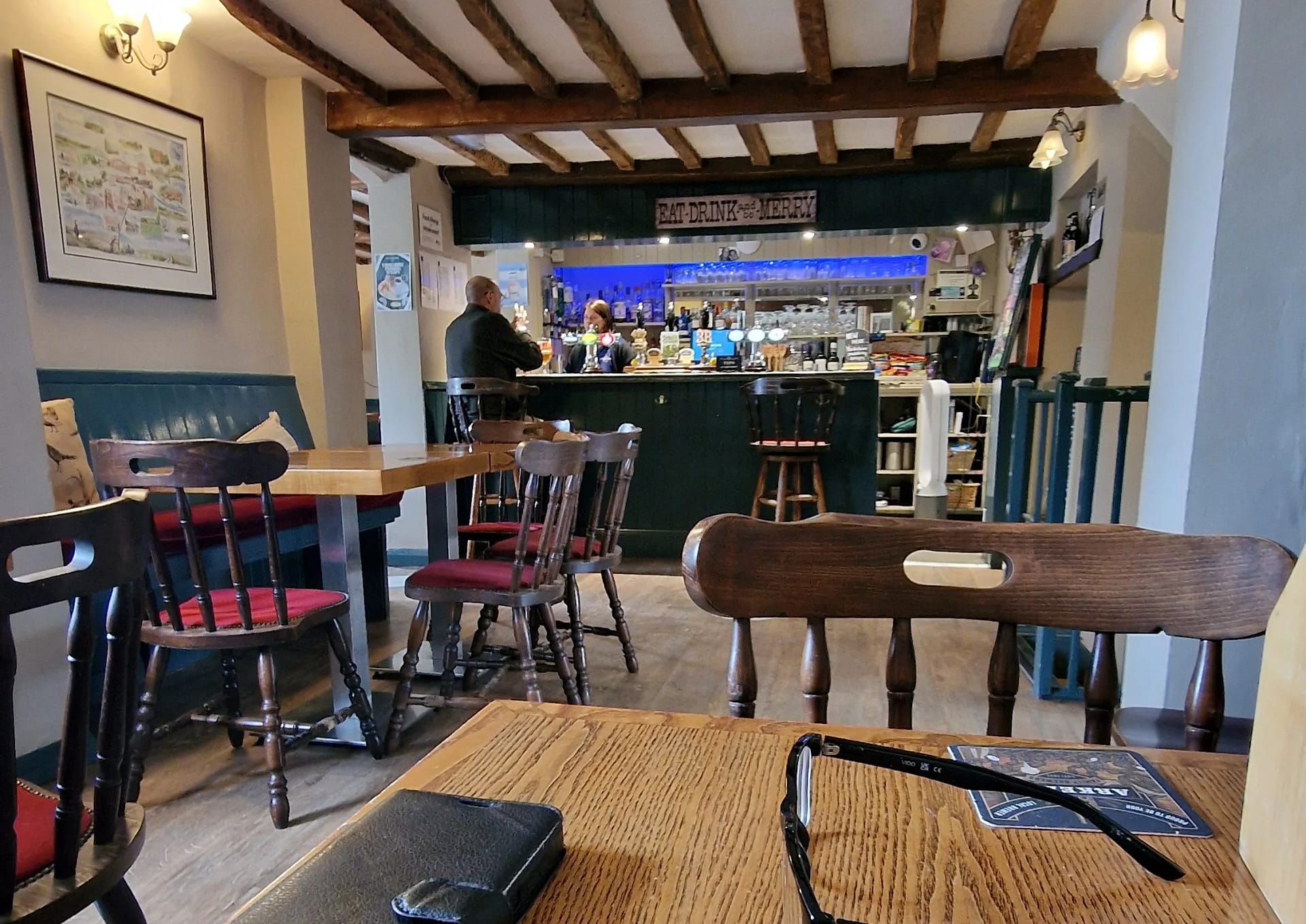 Interior of a traditional English pub with wooden beams, tables, and chairs, showing the bar area where two people are seated and a sign reading “Eat, Drink and Be Merry.”