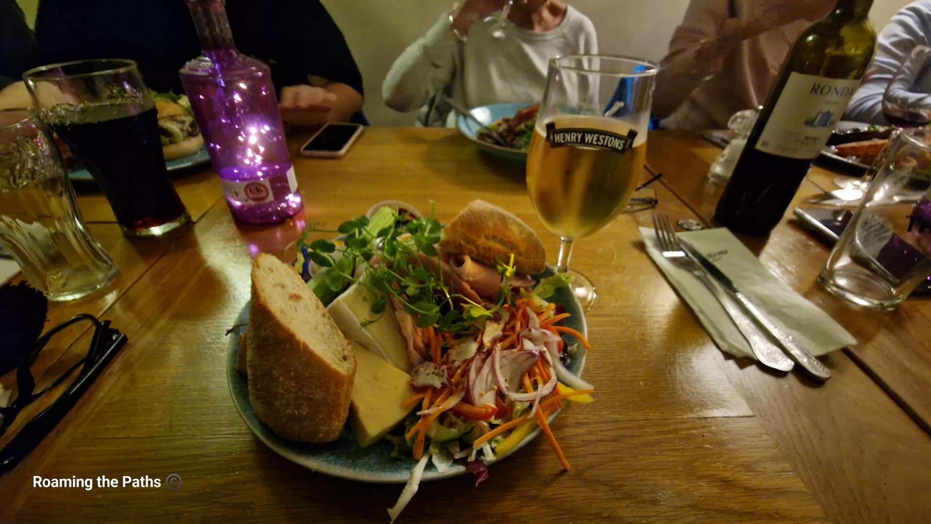 A Ploughman’s lunch served with Henry Westons cider on a wooden pub table, surrounded by friends enjoying drinks and food in a warm, relaxed setting.