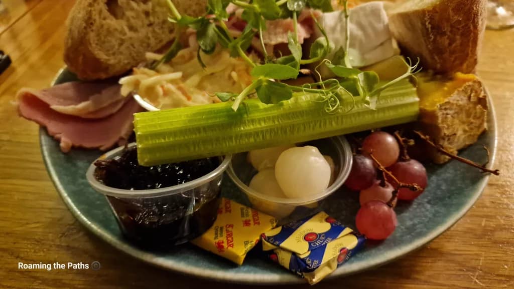 A Ploughman’s lunch served on a rustic plate with slices of ham, cheeses, crusty bread, celery, grapes, pickled onions, Branston pickle, butter portions, coleslaw, and pea shoots for garnish.
