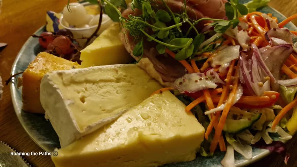 A close-up of a Ploughman’s-style cheese plate with wedges of Somerset Brie, Shropshire Blue, Hereford Hop, and Worcester Gold cheeses, served with grapes, pickled onions, salad, and slices of ham topped with pea shoots.
