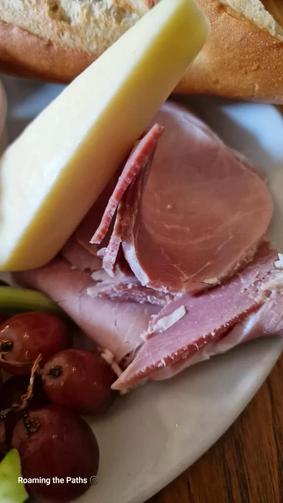 Close-up of sliced ham and a wedge of cheese with red grapes and part of a baguette on a white plate, part of a traditional Ploughman’s lunch.